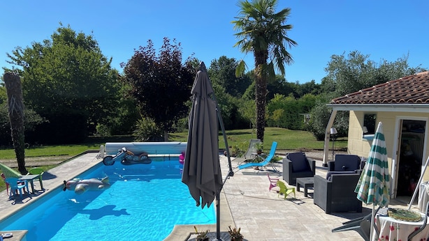 A swimming pool with lounge chairs and umbrellas, surrounded by greenery and a building.