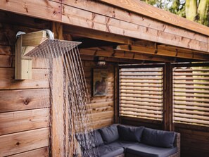 A wooden cabin with a showerhead mounted on the wall, a sofa, and a window with blinds. - Vacation cottage in nature - tiny - peaceful - woods - outdoor sauna (Putten)