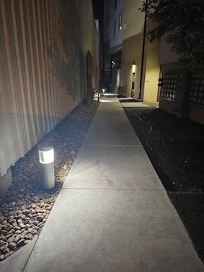 Un chemin illuminé la nuit, bordé de bâtiments, d’une clôture en métal et d’un mur orné de motifs circulaires.