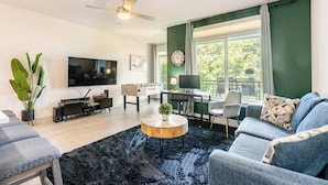 A living room with a flat-screen TV, a coffee table, a sofa, a desk with a chair, and a window with curtains. - Chic Riverwalk Facing Condo Pool View  Free Parking Pets Welcome (San Antonio)