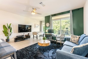 Ein Wohnzimmer mit Flachbildfernseher, Couchtisch, Sofa, Schreibtisch mit Stuhl und einem Fenster mit Vorhängen.