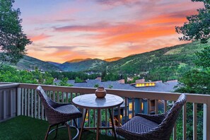 A balcony with wicker chairs and a table, overlooking a valley with houses and trees under a sunset sky. - Steps to Trails + Free Bus to Slopes | Balcony + Views | Serenity by the Slopes (Vail)