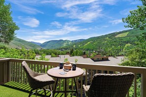 A balcony with wicker chairs and a table, overlooking a valley with houses and mountains. - Steps to Trails + Free Bus to Slopes | Balcony + Views | Serenity by the Slopes (Vail)