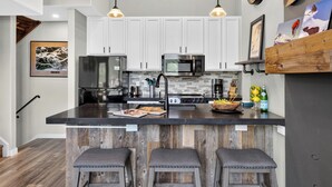 A kitchen with white cabinets, a black countertop, a stainless steel refrigerator, a microwave, and a sink. There are three bar stools and a wooden shelf with decorative items. - Steps to Trails + Free Bus to Slopes | Balcony + Views | Serenity by the Slopes (Vail)