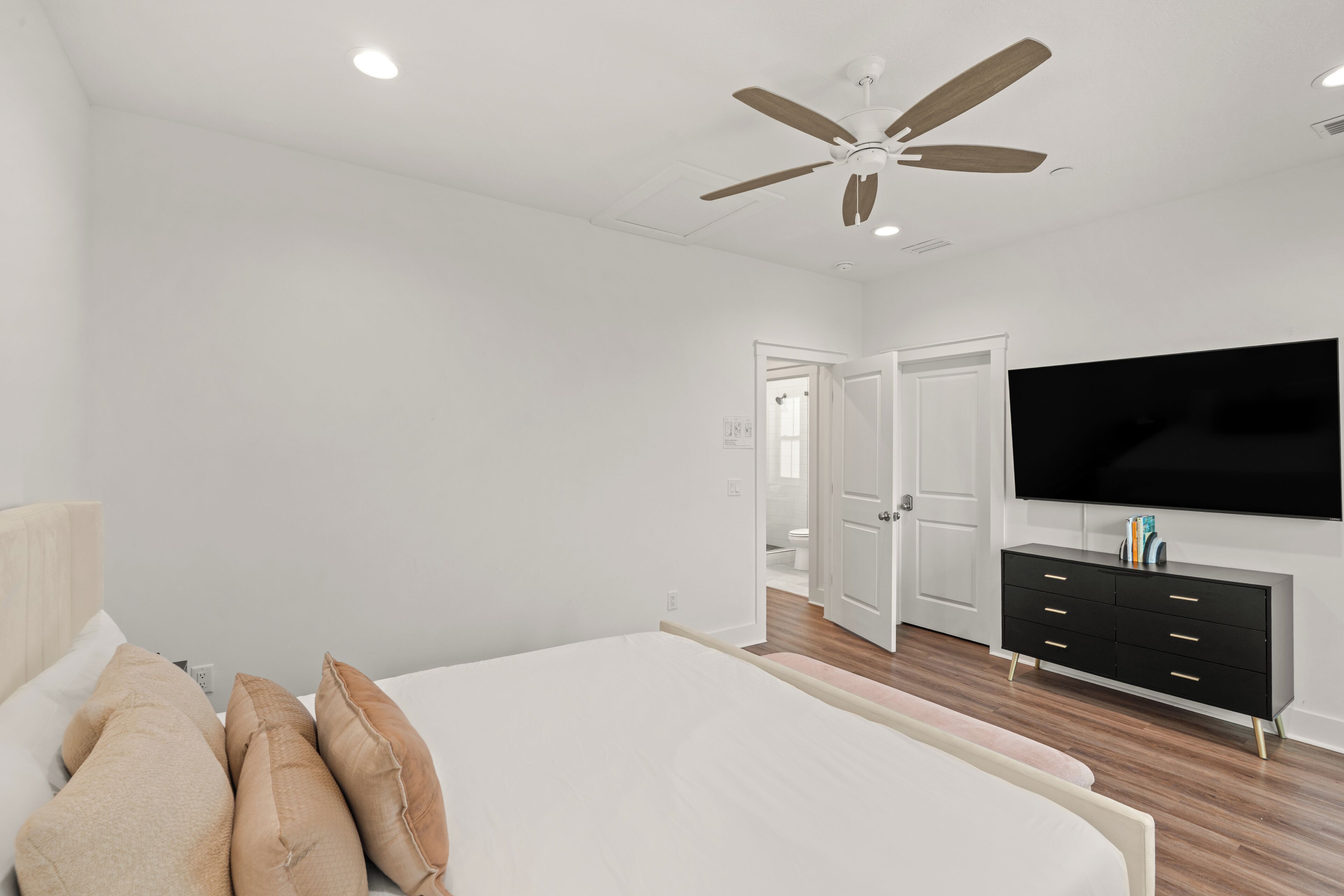 Un dormitorio con cama, ventilador de techo, televisión, cómoda y una puerta que da a un baño.