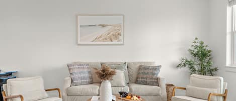 A living room with a sofa, two armchairs, a coffee table, a framed picture, a vase, a glass of wine, and a fruit platter.