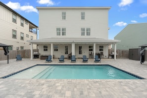 A swimming pool with a covered patio area, outdoor seating, and a large two-story building in the background. - Luxe Beach Retreat • Shared Pool + Hot Tub • Walk to Beach | High Tide (Panama City Beach)