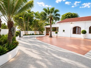 A white building with arched windows and a tiled roof, surrounded by palm trees and a paved courtyard.