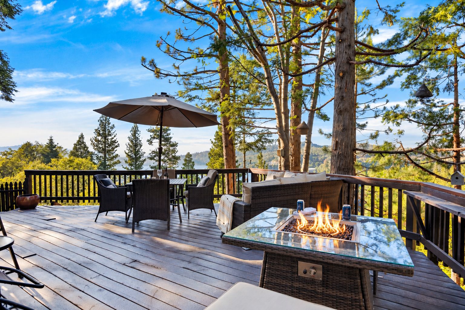 Une terrasse dotée de mobilier d’extérieur, d’une table à plateau en verre avec un foyer intégré, et offrant une vue sur des arbres et une étendue d’eau.