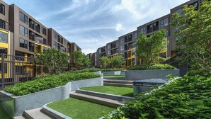 Un moderno edificio residencial con varios balcones, un patio ajardinado con césped artificial y una variedad de árboles y arbustos.