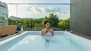 Una persona en un jacuzzi con vistas a un paisaje montañoso, una botella y una silla.
