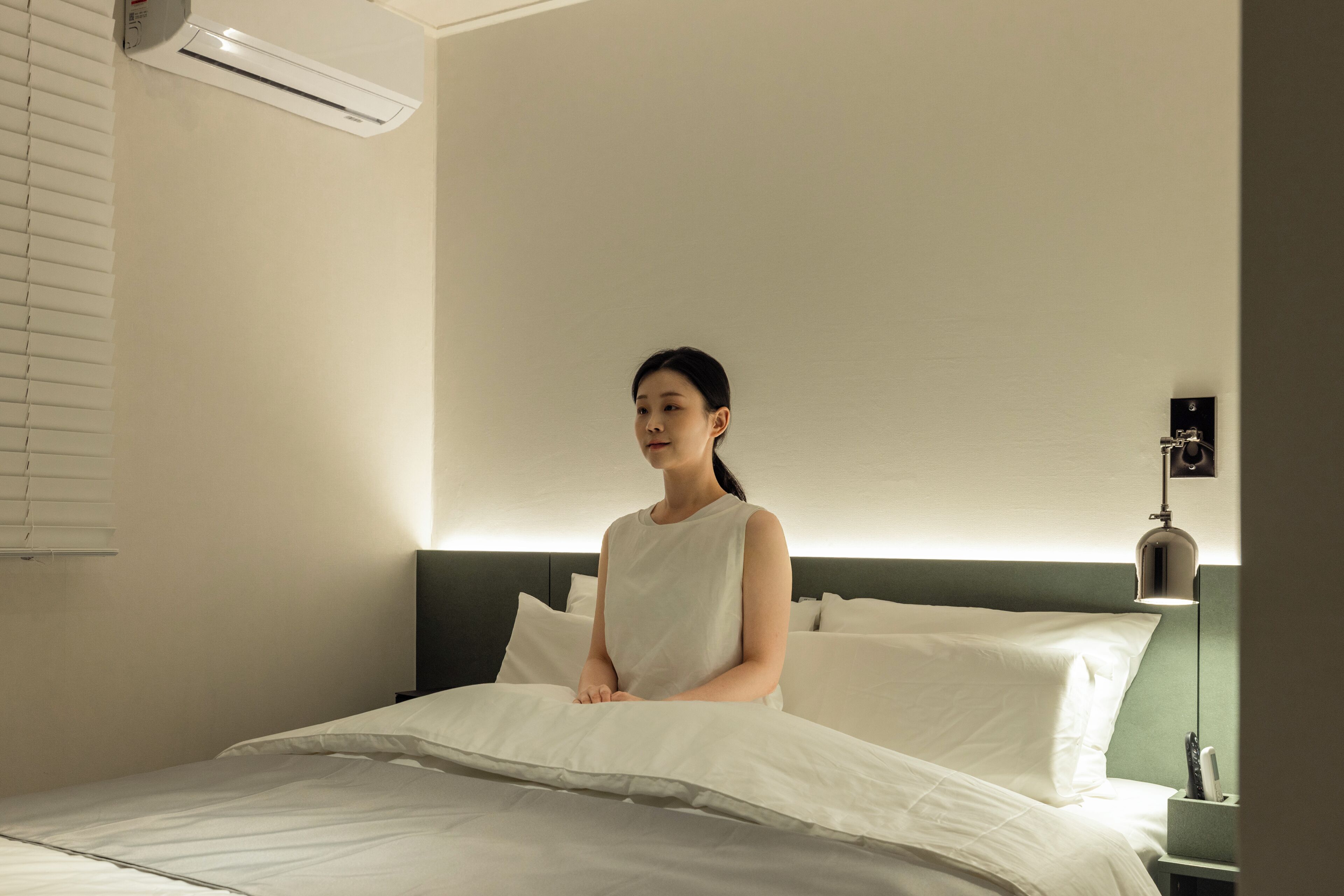 Une personne est assise sur un lit dans une chambre d’hôtel, on peut voir une tête de lit, un appareil de climatisation et une lampe de chevet.