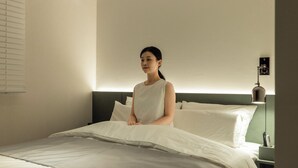 A person sitting on a bed in a hotel room with a headboard, an air conditioning unit, and a bedside lamp.