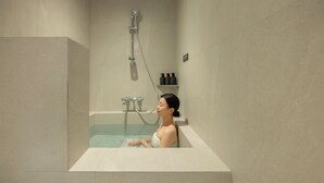 A woman in a bathtub with a showerhead above, a caution sign below, and a shelf with toiletries to the right.