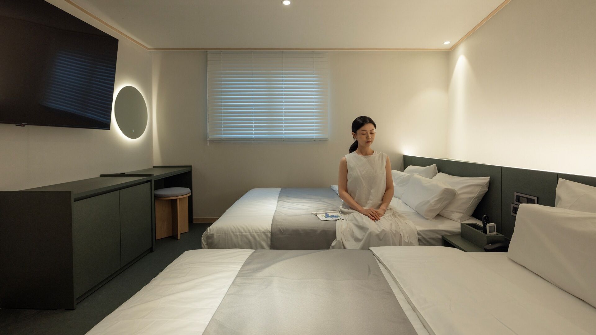 Ein Hotelzimmer mit zwei Betten, einem Schreibtisch, einem Fernseher und einem Fenster mit Jalousien.