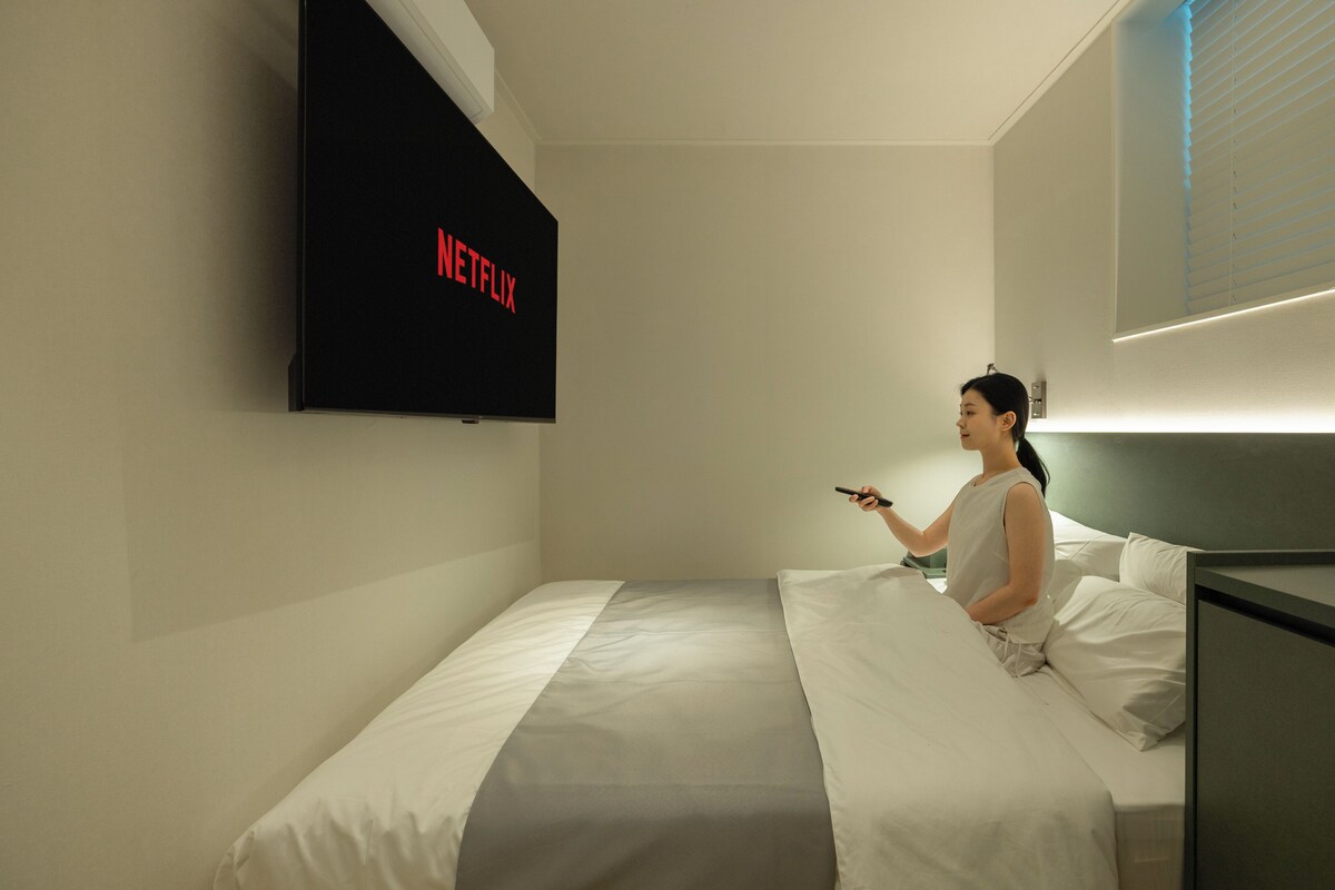 Una habitación de hotel con una cama, un televisor en la pared mostrando 'NETFLIX' y una mesita de noche.