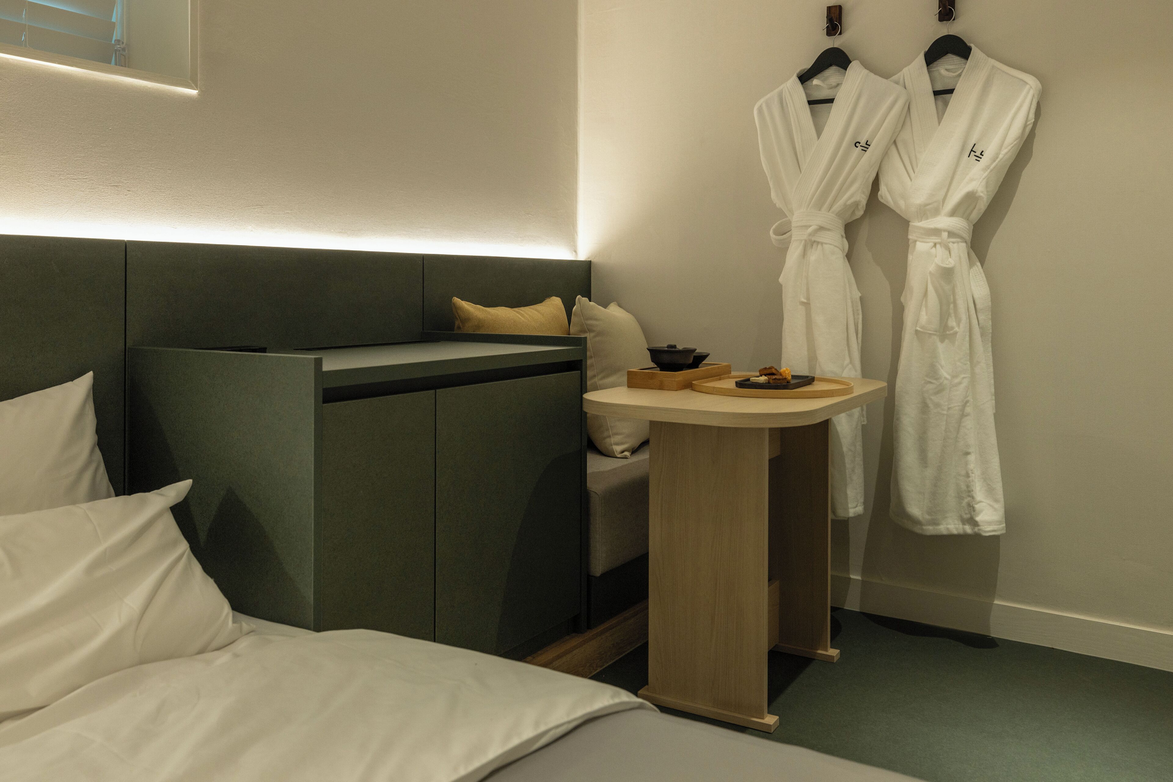 A hotel room with a bed, a sofa bed, a small wooden table, and two white robes hanging on the wall.