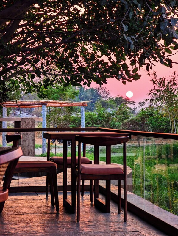 Un área de estar al aire libre con mesas y sillas, una pérgola de madera y vistas al atardecer sobre el agua.