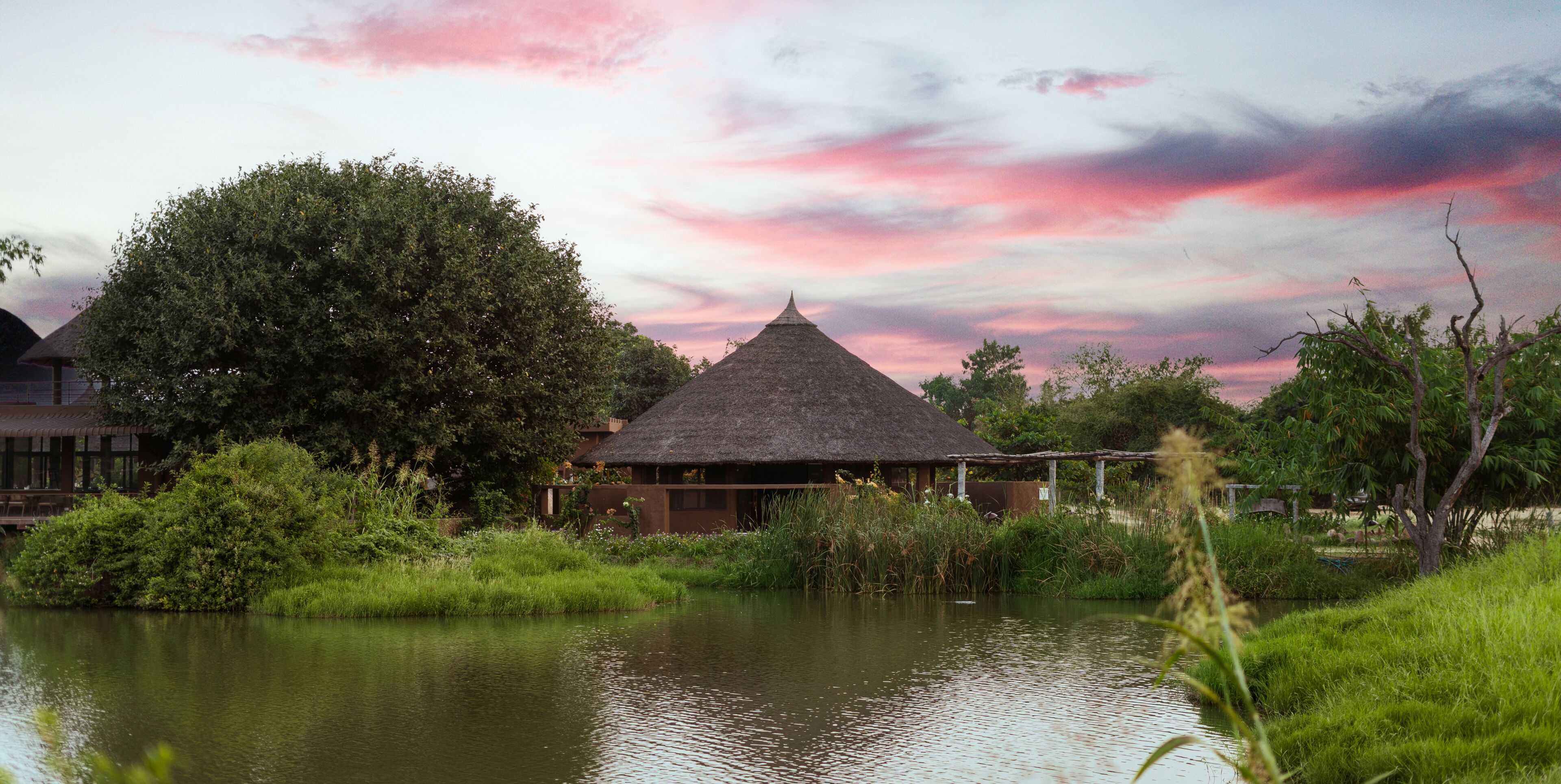 A thatched-roof hut by a calm body of water, surrounded by lush greenery and a clear sky.