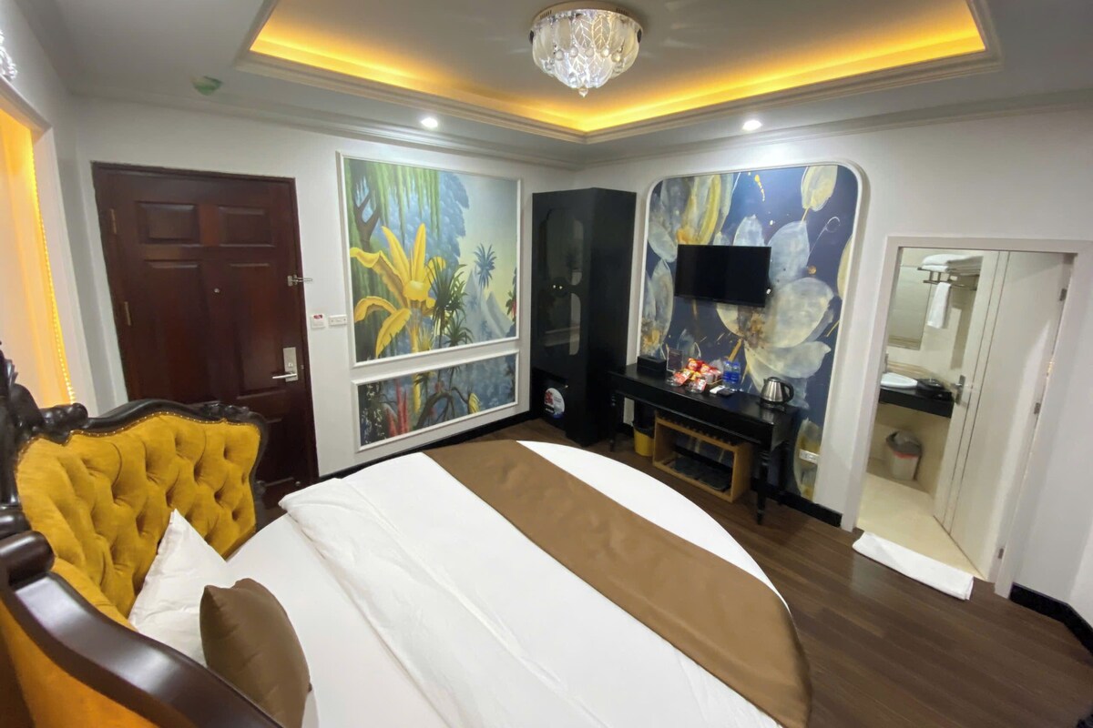 A hotel room with a large bed, a television, a painting, and a mirror.