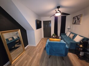 A living room with a blue sofa, a small table with a plant, a ceiling fan, and a large mirror.
