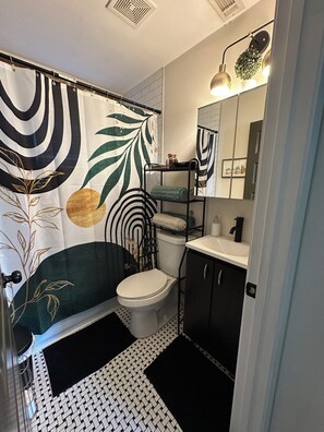 Un baño con cortina de ducha de estampado blanco y negro, inodoro blanco, lavamanos negro y espejo con una planta.