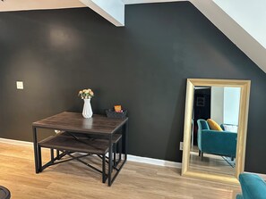 A room with a dark wall, a wooden table with a vase and snacks, a mirror with a gold frame, and a teal chair.