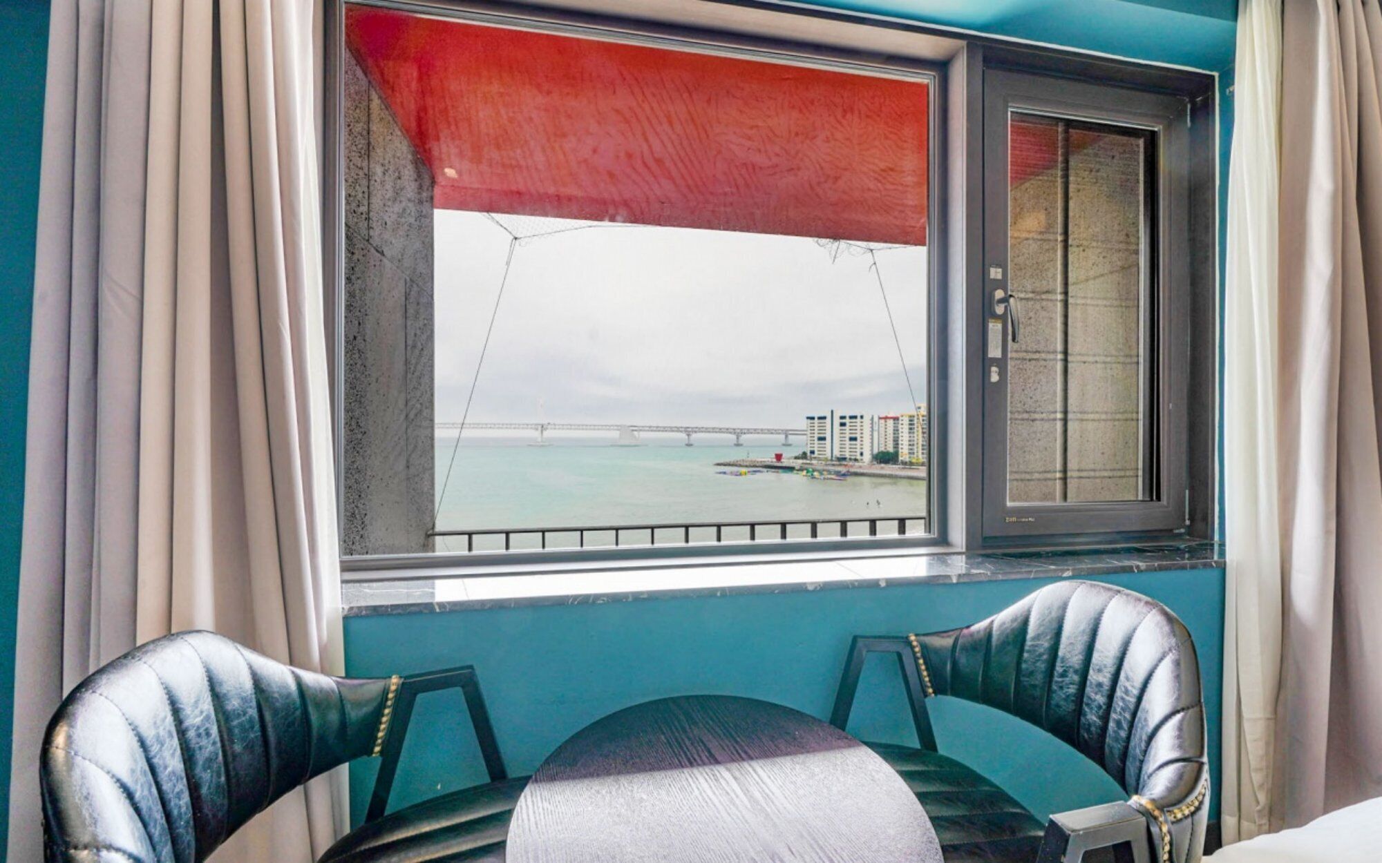 Una habitación con vistas a un puente y edificios al otro lado del agua, una ventana con persiana roja y dos sillones de cuero.