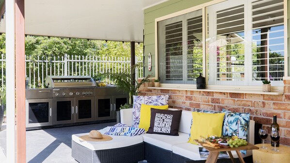 An outdoor seating area with a grill, cushioned seating, and a window with shutters.