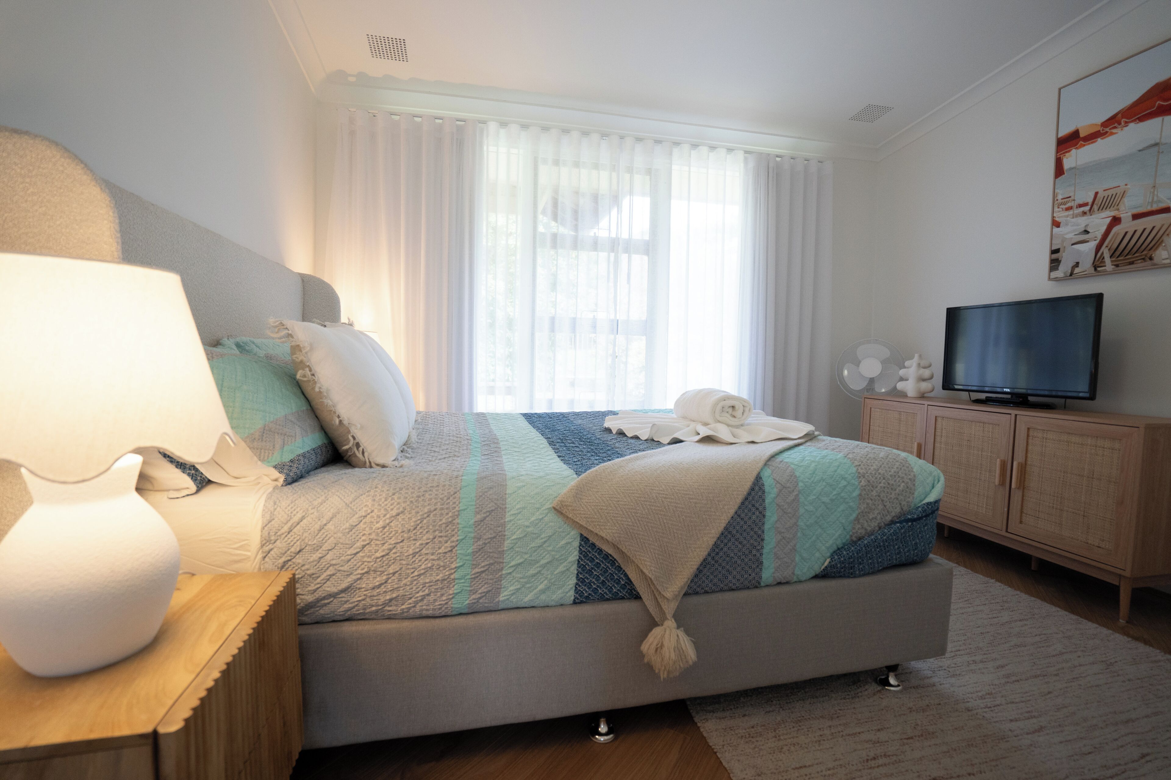 A bedroom with a bed, a bedside table with a lamp, a television, and a window with curtains.