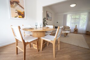 A dining area with a round wooden table, white chairs, and a large window with sheer curtains. - Self contained villa, Middleton Rd Cottage (Mira Mar)