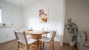 Et spiseområde med et rundt bord, fire stole, en potteplante og en hvid væg med et indrammet billede.