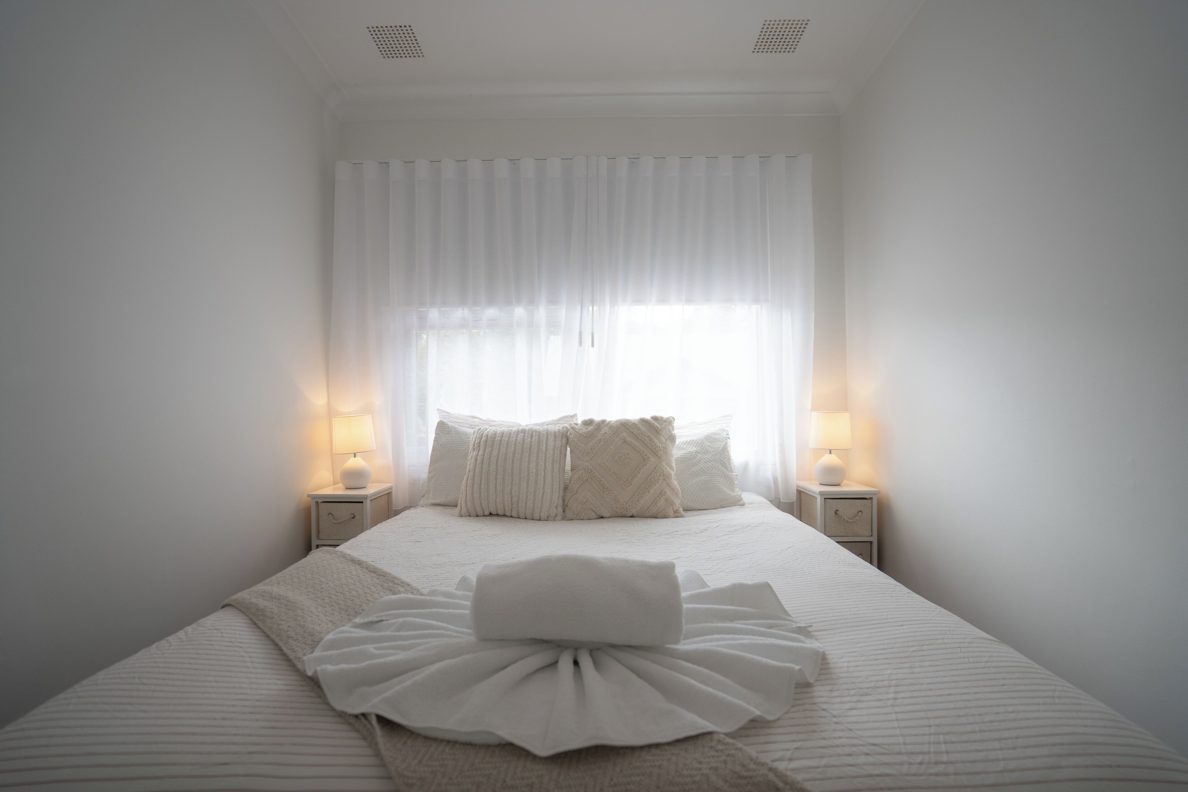 A neatly made bed with a white duvet and pillows, two bedside lamps, and a window with sheer curtains.