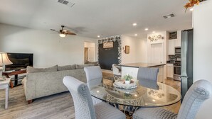Ein modernes Wohnzimmer mit Glastisch, grau gepolsterten Sesseln, einer Couch, einem Deckenventilator und einer Küche im Hintergrund.