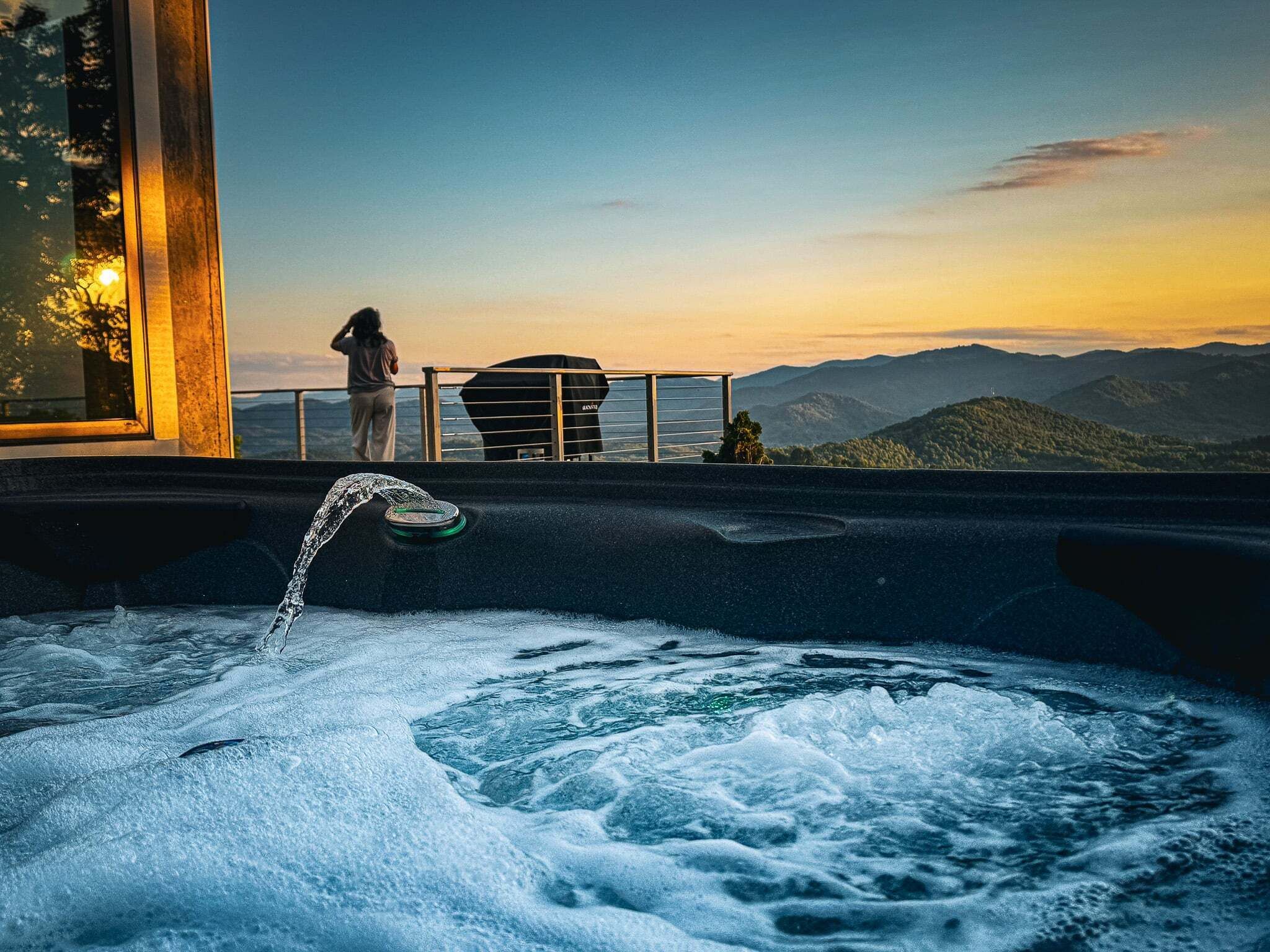 Un bain à remous avec de l’eau en mouvement, une personne qui se tient à proximité, et une vue sur les montagnes au coucher du soleil.