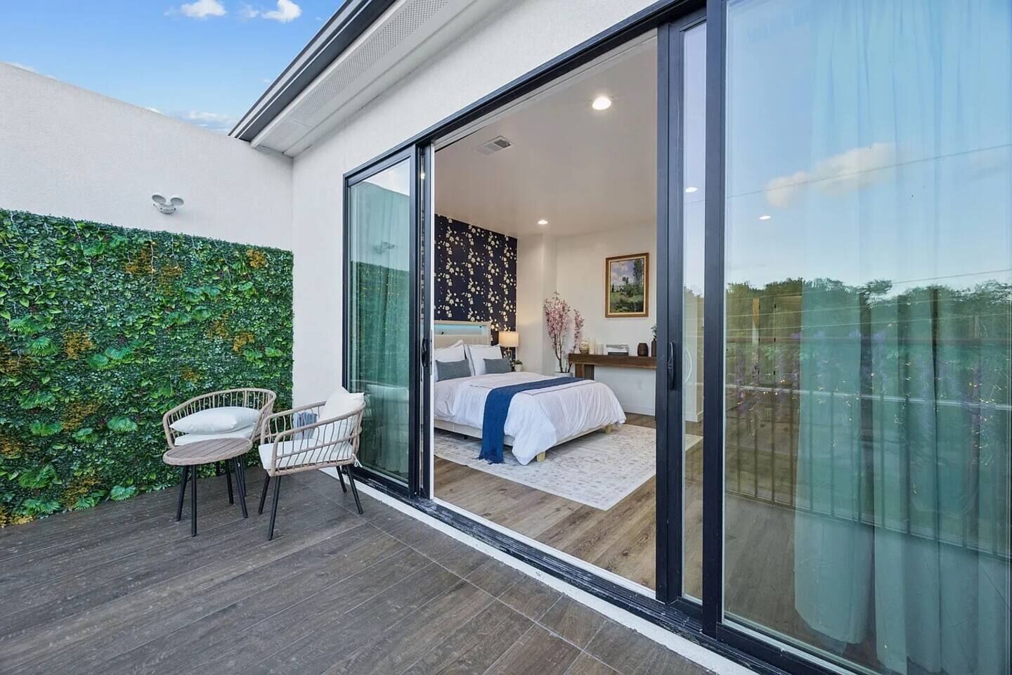 Un dormitorio con una cama, una silla, un escritorio y un amplio ventanal corredero que da a un balcón con un muro verde.