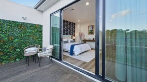 Et soveværelse med seng, stol, skrivebord og en stor skydedør, der fører ud til en balkon med en grøn væg.