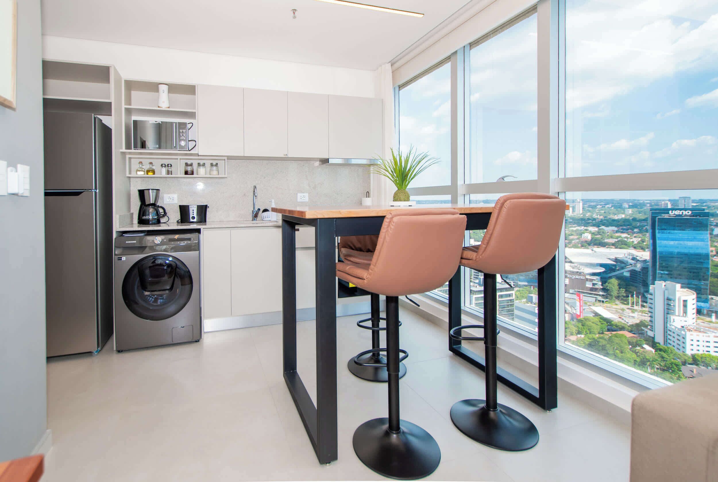 Une cuisine moderne équipée d’un lave-linge, d’une machine à café et d’une grande fenêtre donnant sur la ville.