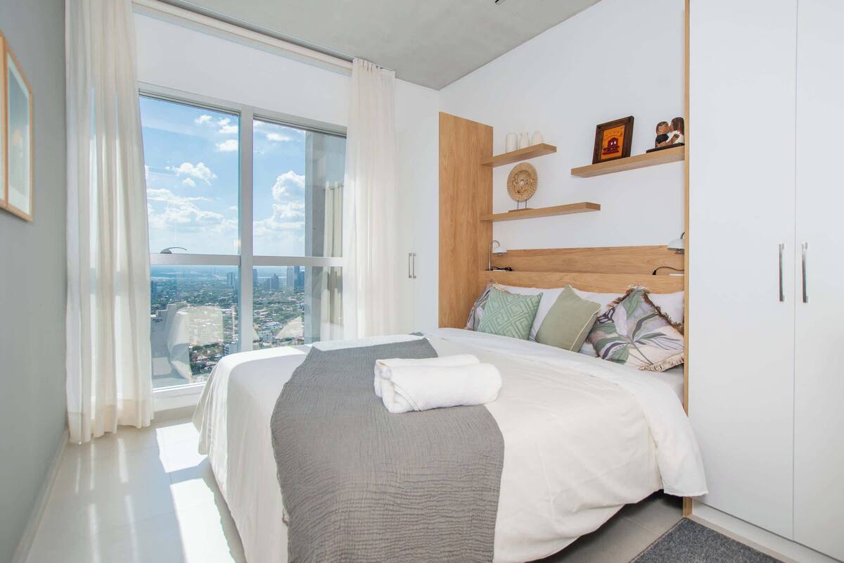 Ein modernes Schlafzimmer mit großem Fenster, einem Bett mit weißer und grauer Decke, Holz-Kopfteil und Blick auf die Stadt.
