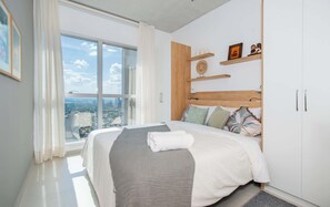 Ein modernes Schlafzimmer mit großem Fenster, einem Bett mit weißer und grauer Decke, Holz-Kopfteil und Blick auf die Stadt.