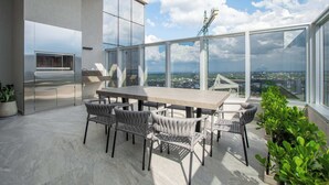 Un área de comedor exterior moderna con una mesa grande y sillas, una parrilla incorporada y vistas de la ciudad.