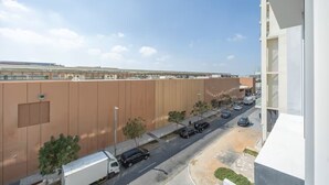 A view of a street with a large brown building, a white building with balconies, and a clear sky. - Bloomfields Studio - Oasis 1 - Masdar (Abu Dhabi)