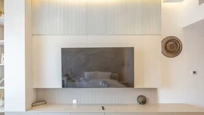 A modern living room with a large flat-screen TV mounted on a white wall, a minimalist console table below, and a decorative wall piece to the right. - Bloomfields Studio - Oasis 1 - Masdar (Abu Dhabi)
