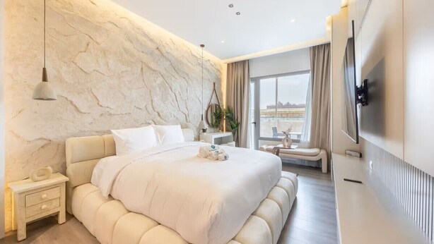 A modern bedroom with a large bed, a TV, a nightstand, and a view of the city. - Bloomfields Studio - Oasis 1 - Masdar (Abu Dhabi)