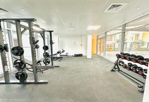 Ein Fitnessstudio mit Krafttrainingsgeräten, darunter Hanteln und Kurzhanteln, und großen Fenstern mit Blick ins Freie.