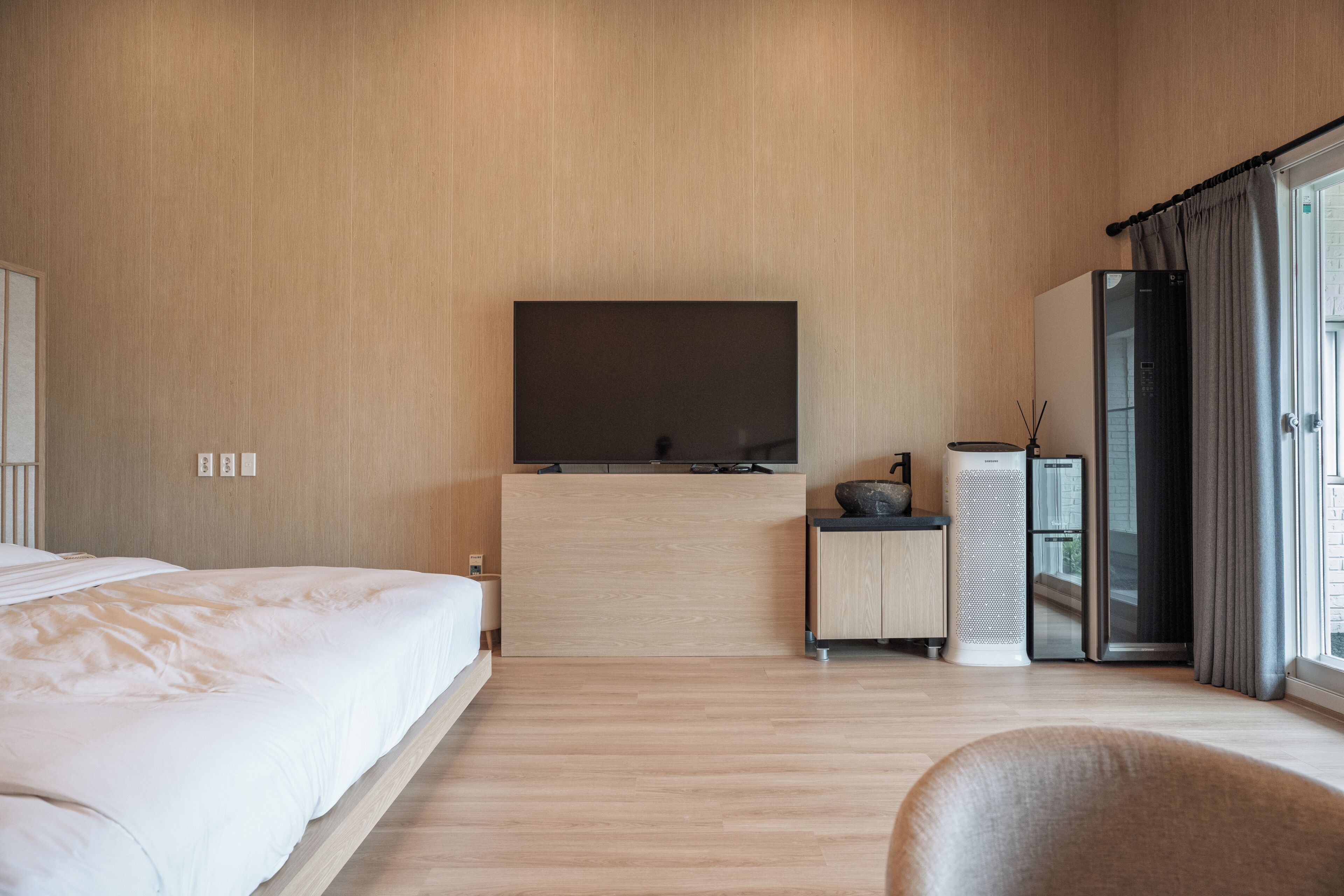 A hotel room with a bed, a flat-screen TV mounted on the wall, a minibar, a refrigerator, and a window with curtains.