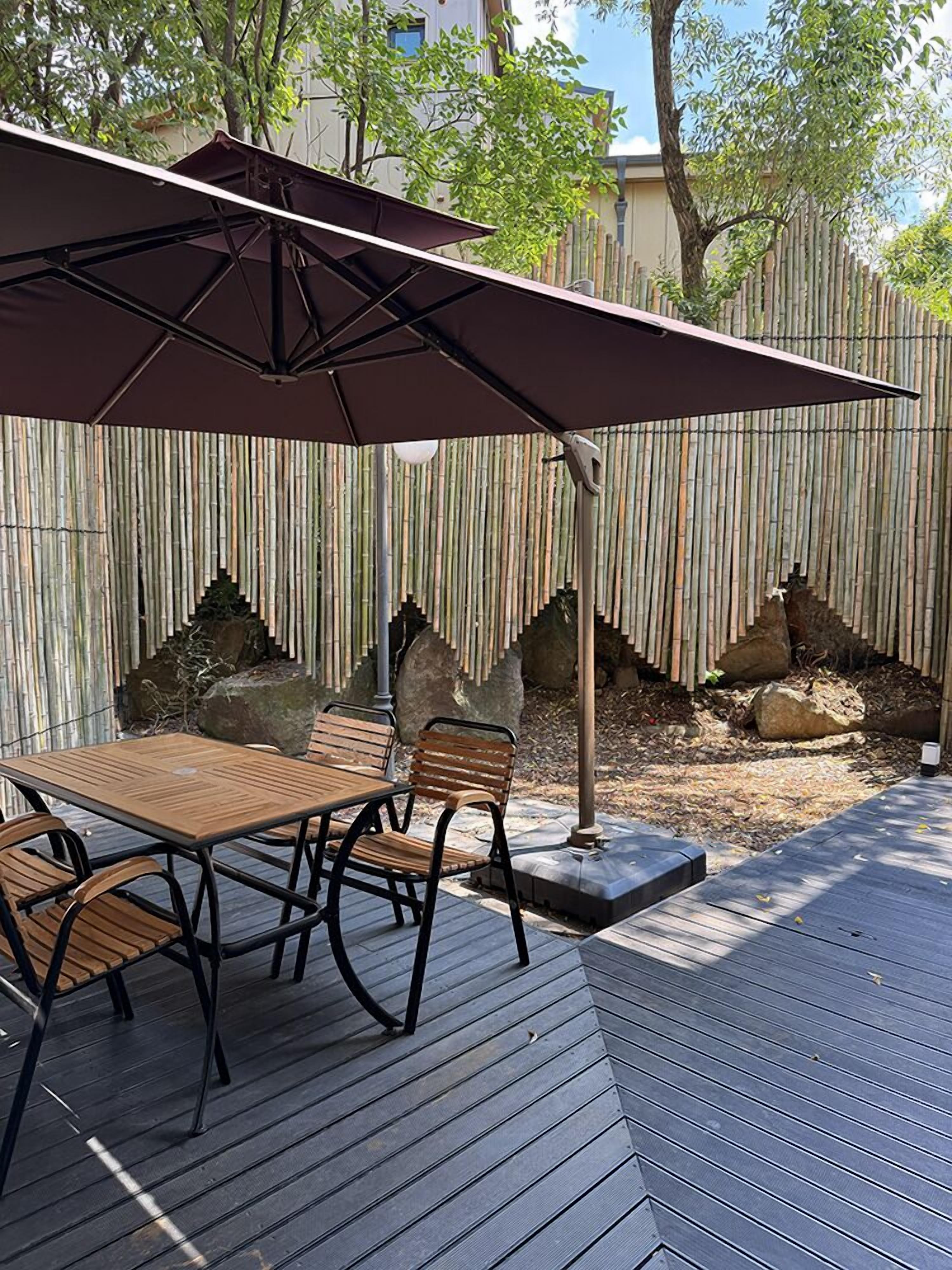 Un patio al aire libre con una mesa, sillas, un paraguas grande y una valla de bambú al fondo.