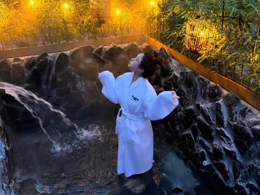 A person in a white robe standing in a hot spring with a waterfall backdrop and warm lighting.
