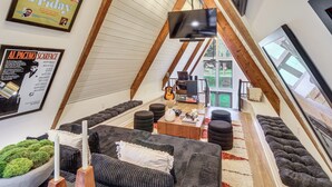A loft-style room with a sloped ceiling, a flat-screen TV mounted on the wall, a wooden coffee table, and a seating area with dark sofas and ottomans. - Hot Tub, Grill & Fire Pit: Quiet Georgetown Escape (Savannah)