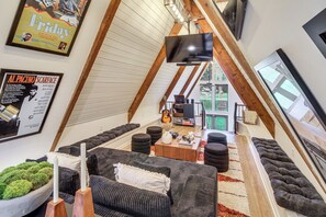 A loft-style room with a sloped ceiling, a flat-screen TV mounted on the wall, a wooden coffee table, and a seating area with dark sofas and ottomans. - Hot Tub, Grill & Fire Pit: Quiet Georgetown Escape (Savannah)
